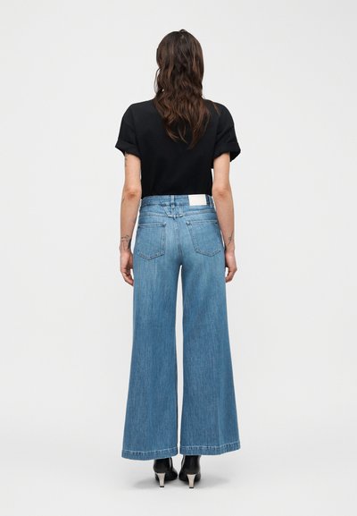 Άτομο με μακριά μαλλιά που φοράει μαύρο τοπ με κοντά μανίκια, τζιν παντελόνι ψηλόμεσο, φαρδύ και μπλε, καθώς και μαύρα παπούτσια με ψηλά τακούνια, απεικονιζόμενο από πίσω.