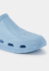 Sapato azul-claro em estilo slip-on, feito de material semelhante a borracha. Apresenta orifícios de ventilação e detalhes texturizados nas laterais para maior aderência e respirabilidade.