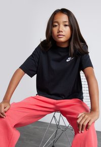 Camiseta negra de algodón con un logo blanco de Nike, combinada con pantalones de chándal coral de corte holgado, sentado en una silla de alambre metálico.