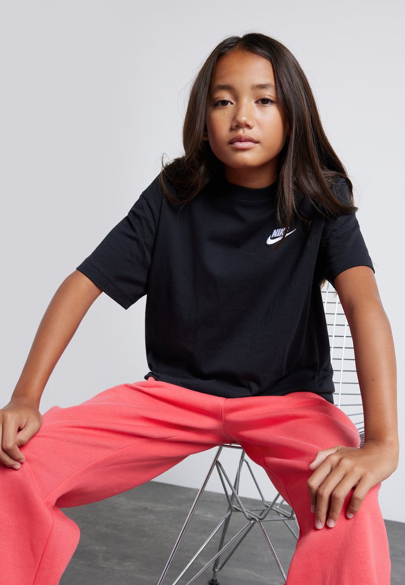 Camiseta negra de algodón con un logo blanco de Nike, combinada con pantalones de chándal coral de corte holgado, sentado en una silla de alambre metálico.