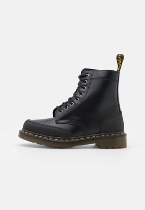 Botte basse montante en cuir noir à lacets avec couture jaune, bout renforcé et semelle en caoutchouc translucide, vue de profil sur fond blanc.