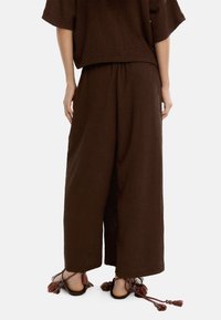 Brązowe lniane spodnie o szerokich nogawkach z elastycznym paskiem w talii, w połączeniu z sandałami z wielokolorowymi frędzlami i plecionymi paskami.