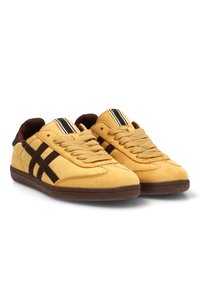 Zapatillas de ante amarillas con acentos marrones y bandas decorativas negras. Presentan una puntera redonda y un diseño de cordones en la parte delantera.