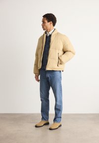 Jachetă puffer bej cu manșete ribbate și guler în picioare, purtată peste o cămașă din denim închis, asortată cu blugi albastru deschis și cizme de culoare crem.