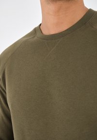 Sudadera verde oliva con cuello redondo acanalado, textura suave y mangas raglán. Presenta detalles de costura mínimos en los hombros.