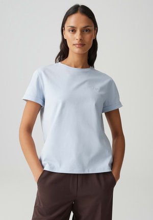 Donna che indossa una t-shirt a maniche corte azzurro chiaro con risvoltini e pantaloni marrone scuro, in piedi con le mani in tasca davanti a uno sfondo semplice.