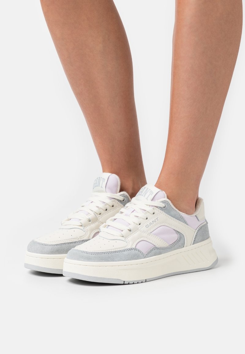 Paire de baskets de couleur claire avec des lacets blancs et des accents gris portées sur des jambes nues sur un fond blanc uni.