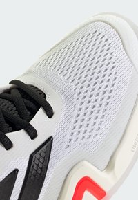 Λευκό αθλητικό παπούτσι με υφαντό πλέγμα στο επάνω μέρος, μαύρους κορδόνια και γκρι λεπτομέρεια. Διαθέτει κόκκινη λεπτομέρεια στη φτέρνα και ελαφρύ πάτο.