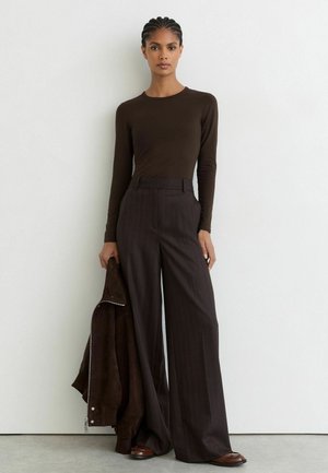 Frau mit geflochtenem Haar, die ein dunkelbraunes Langarmshirt und weit geschnittene Hosen trägt und eine passende Jacke vor einem schlichten Hintergrund hält.