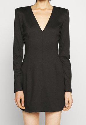Femme portant une mini-robe noire cintrée à manches longues, avec un décolleté en V profond et des épaules structurées, sur un fond uni.