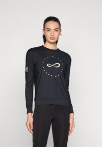 Svart langermet topp med rund hals, med et sentralt uendelighetssymbol design og merket ermelapp. Sammen med svarte leggings.