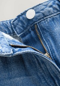 Primer plano de unos jeans de mezclilla azules con un cierre de cremalleras de latón parcialmente desabrochado y un botón plateado en la cinturilla.