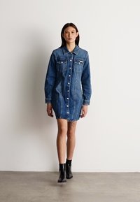 Λιντζάκι τζιν σε μεσαίο μπλε χρώμα. Διαθέτει κουμπωτή μπροστινή ανοιχτή, δύο τσέπες στο στήθος, μακριά μανίκια και ελαφρώς στενεμένη hem. Φοριέται με μαύρες μπότες μέχρι τον αστράγαλο.