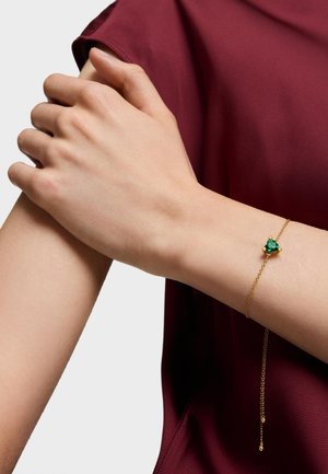 Pulsera de cadena de oro con una piedra verde central, llevada en un brazo de tono de piel claro. El fondo presenta una tela de burdeos profundo.