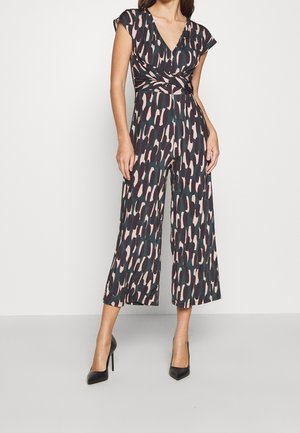 Kvinde iført en sort jumpsuit med pink og grøn abstrakt mønster, korte ærmer, V-hals, vide ben og sorte højhælede sko.