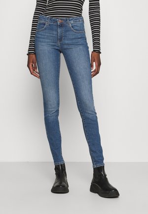 Kobieta w obcisłych niebieskich dżinsach, czarnych botkach za kostkę i czarno-białej, długorękawowej bluzce w paski, stojąca na tle jednolitego tła.