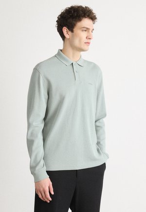 Light green long-sleeve polo shirt made of ribbed textured fabric with a three-button placket and subtle logo. Paired with black pants.