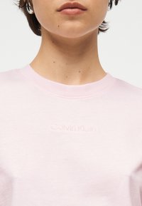 Γυναίκα που φοράει ανοιχτό ροζ μπλούζα με στρογγυλό λαιμό και ανάγλυφο λογότυπο Calvin Klein, ασημένια σκουλαρίκια κρίκους και κοντά καστανά μαλλιά.