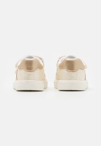 Beige tenisky s texturovaným materiálem a lesklými akcenty, s bílou podešví a vlnkovým designem na patě. Zobrazeno z zadního pohledu.