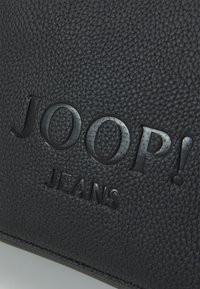 Čierny textúrovaný kožený produkt s embosovaným brandingom "JOOP! JEANS". Povrch má charakteristickú štruktúru a matný povrch.