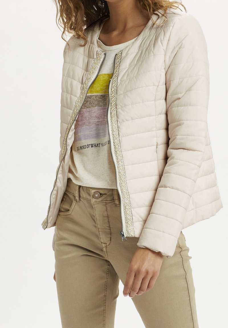 Jasnoróżowa pikowana kurtka z zamkiem z przodu i teksturowanym srebrnym wykończeniem. Nosi się ją na beżowym T-shircie z grafiką i beżowych spodniach, z bocznymi kieszeniami.