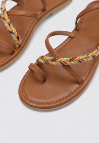 Sandalias de cuero marrón con tiras trenzadas de tejido multicolor, suelas planas y diseño minimalista.