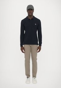 Camisa polo de manga longa azul-marinho com pequeno logotipo, calças bege slim-fit, boné cinza e ténis brancos com atacadores estampados. Design minimalista.