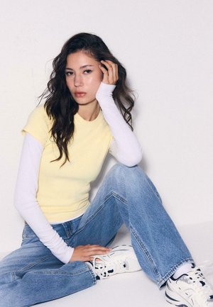 Ung kvinde med langt mørkt hår, siddende med benene krydsede, iført lagdelte gule og hvide toppe, blå jeans og hvide sneakers mod en ensfarvet baggrund.