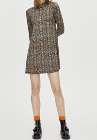 Robe à manches longues avec un motif complexe marron et beige, dotée d'un col montant et d'un ourlet évasé. Associée à des chaussettes orange et des bottes noires.