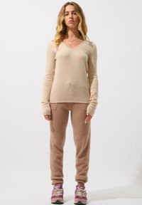 Beige kashmirtröja med V-ringning; bruna ulljoggers med framfickor; modellen bär rosa sneakers med grå sulor och snören.