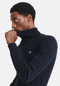 Pulover tricotat de culoare bleumarin cu guler înalt, având un model cu cabluri, manșete și guler cu nervuri, cu un logo mic pe piept. Textură moale.