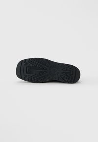 Semelle en caoutchouc noir d'une chaussure avec des motifs de bande de roulement géométriques et un logo circulaire au centre, sur un fond clair uni.