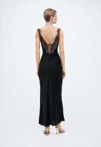 Γυναίκα που φοράει μαύρο αμάνικο φόρεμα με δαντελένιες λεπτομέρειες και βαθιά V στο πίσω μέρος, στέκεται με γυμνά ψηλοτάκουνα παπούτσια μπροστά σε απλό λευκό φόντο.