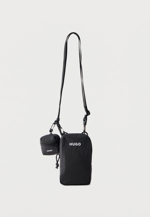Fekete keresztpántos táska és kisebb pénztárca, mindkettőn fehér "HUGO" logóval, állítható pánttal és cipzáras záródással, egyszerű háttér előtt lógva.
