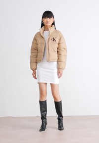 Femme portant une doudoune beige, une robe blanche et des bottes noires montantes contre un fond blanc uni.