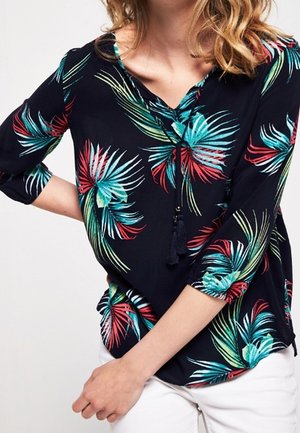 Frau trägt eine marineblaue Bluse mit roten, blaugrünen und grünen tropischen Blattmustern und weiße Hosen, posiert mit einer Hand in der Hüfte.