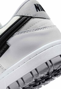 Zapatilla Nike con parte superior de cuero blanco liso, acentos negros, suela texturizada y costuras reforzadas. Cuenta con un logo distintivo en el lateral.