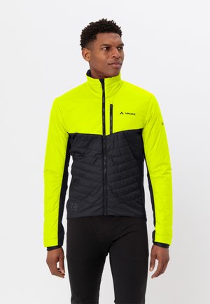 Mann in einer leuchtend gelben und schwarzen Radjacke mit Reißverschluss, der vor einem schlichten weißen Hintergrund steht und nach links schaut.