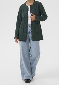 Zelená texturovaná bunda se žebrovaným vzorem, dvěma předními kapsami a řaseným lemem. Kombinována se světle modrými džíny s širokými nohavicemi a černými botami.