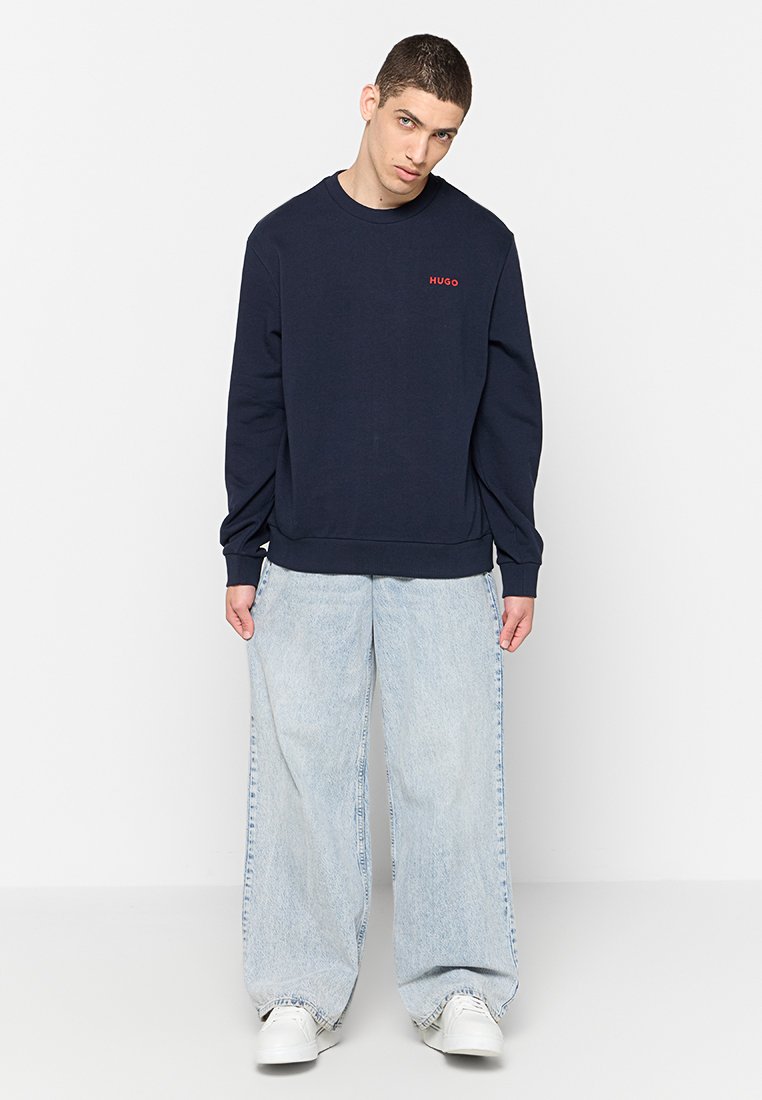 Dunkelblaues Sweatshirt mit Rundhalsausschnitt und rotem Logo auf der Brust, kombiniert mit hellblauen, weit geschnittenen Jeans und weißen Sneakers.