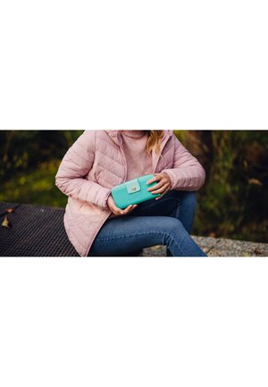 Turquoise wallet with a smooth texture and a gold button, held in a hand, paired with a pink quilted jacket and blue jeans.