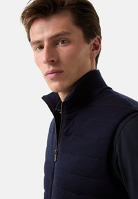 Granatowa kamizelka z wysokim kołnierzem i zamkiem błyskawicznym, z pikowanym wzorem, teksturowanym materiałem i warstwowym wyglądem. Zbliżenie na obszar barku.