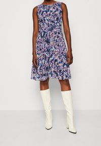 Robe fleurie avec un corsage ajusté, une jupe évasée et des motifs violets, bleus et roses. Associée à des bottes blanches hautes jusqu'aux genoux.