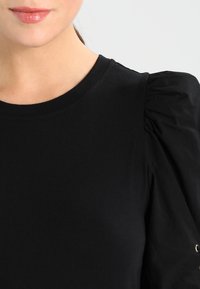 Primo piano di una donna che indossa un top nero con maniche a sbuffo e labbra lucide, mostrata dalle spalle a poco sotto il mento.