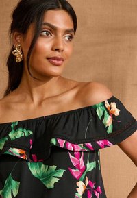Top nero off-shoulder con stampa floreale colorata, scollatura a volant e rifiniture in pizzo decorativo. Orecchini statement dorati completano il look.