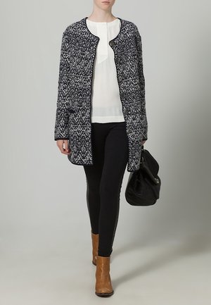 Femme marchant, portant un manteau à motifs noirs et blancs, un chemisier blanc, un pantalon noir, des bottines marron, et portant un sac à main noir.