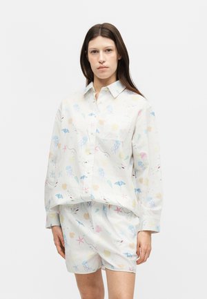 Žena nosiaca bielu košeľu na gombíky a zodpovedajúce šortky s pastelovými motívmi morského života, stojaca pred jednoduchým bielym pozadím.