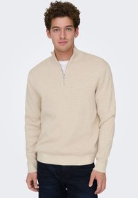 Beige zip-up sweater in soft fabric with long sleeves and a fitted waist. Features a small, metal zipper at the neckline for added detail.