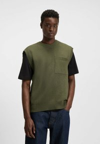 Grüner, gestrickter ärmelloser Pullover mit geripptem Kragen und einer Fronttasche, über einem kurzärmligen schwarzen Shirt getragen, kombiniert mit blauen Jeans.