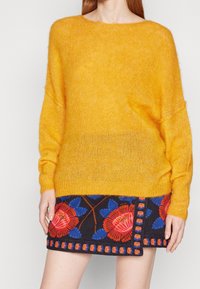 Gul fluffig tröja med lång ärm, matchad med en mönstrad marinblå kjol som har stora röda och blå blommor broderade samt kontrasterande sömmar.